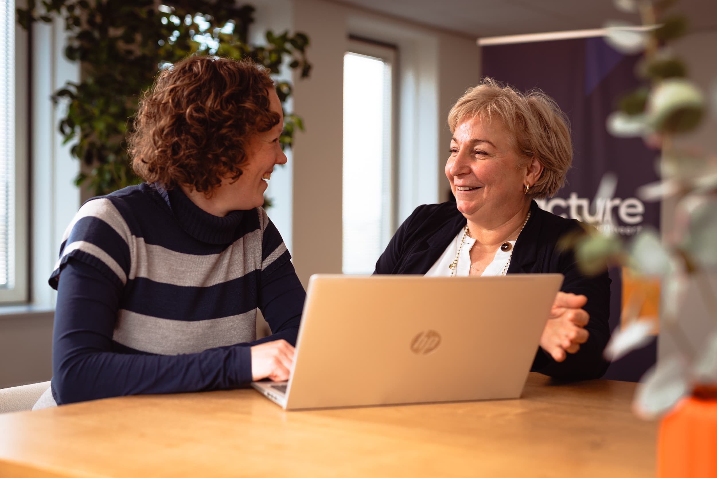 Twee vrouwelijke collega's zitten achter een laptop en lachen naar elkaar.