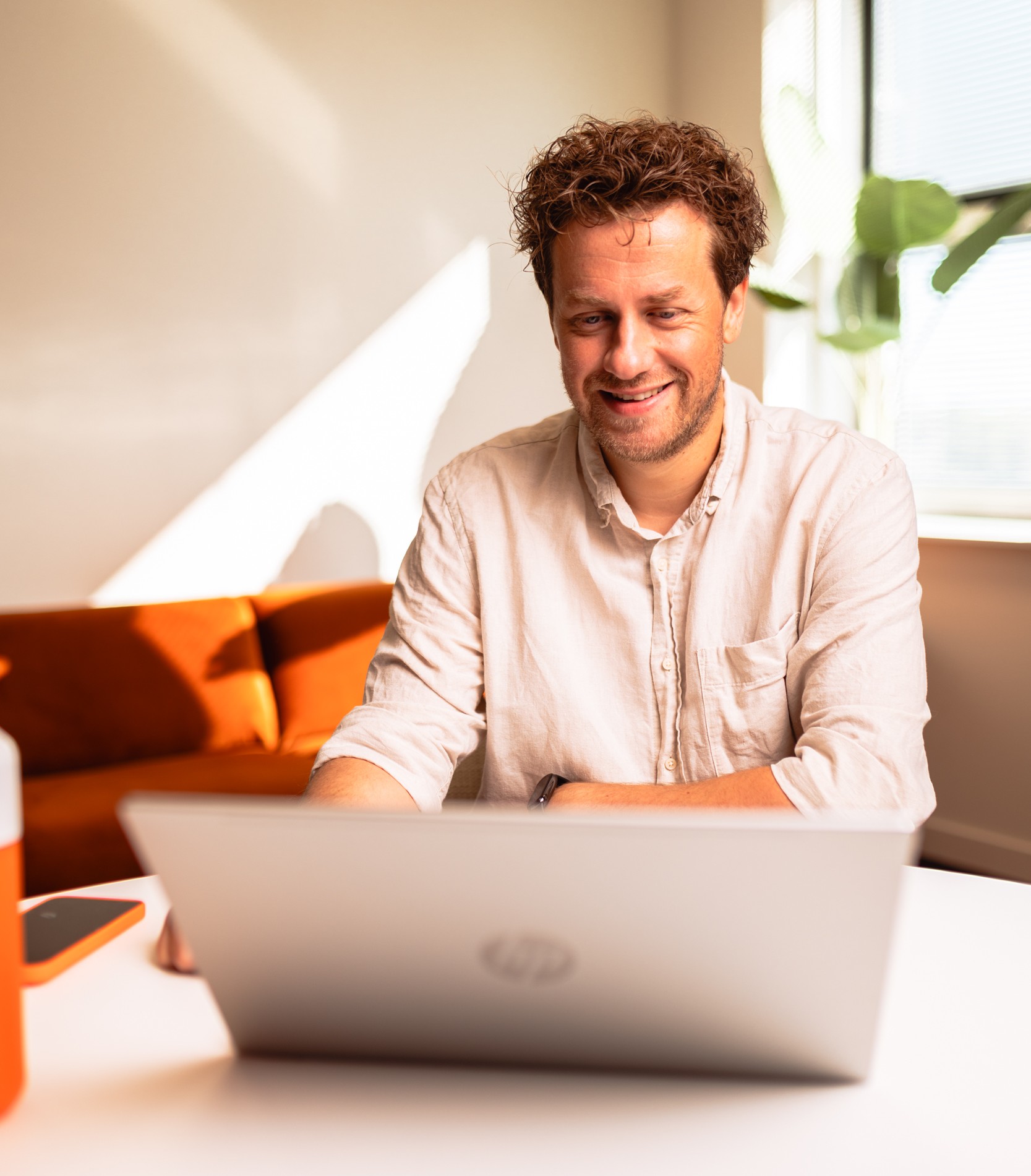 Collega achter computer, lacht en zit in een woonkamer achtige omgeving.